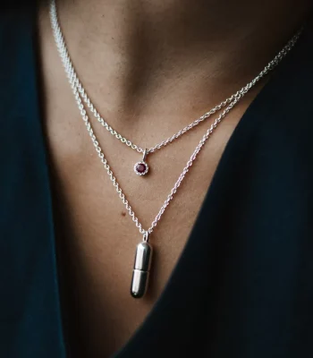 Image of a models neck wearing two silver chains, one with a silver capsule pendant and one with a ruby birthstone pendant.