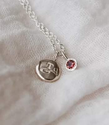Close up of silver chain with silver impression pendant and Pink Tourmaline birthstone pendant on a silver cable chain. The necklace is resting on a piece of white gauze.