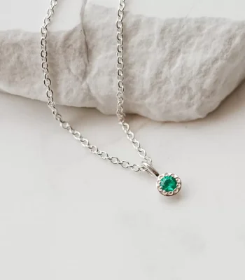Flat lay image of a silver natural emerald birthstone pendant on a silver cable chain. The necklace is draped over a slab of white marble.