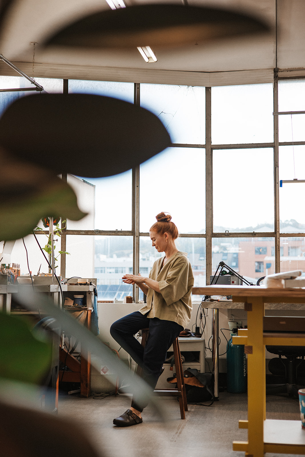 Image of GG sitting on a stool by the window in her Wellington studio, surrounded by jewellery benches and plants. Behind her, the studio features ceiling-high windows looking out to neighbouring buildings.