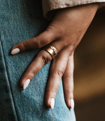 Image of a models hand resting on her leg. She is wearing blue jeans and has pink painted nails. She wears a yellow gold cigar ring on her ring finger. The ring is smooth on one half and roughly textured on the other half.